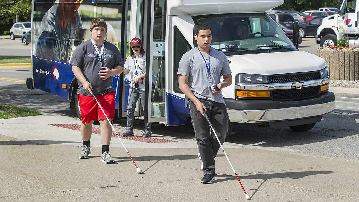 Individuals are navigating outside with white canes while a bus is parked nearby.