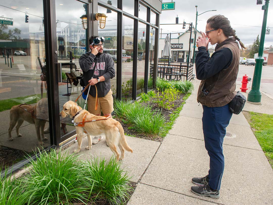 A person stands outside a café, taking a photo while another person and a service dog look into the window.