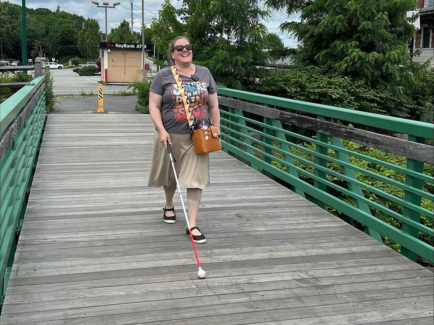 A person walks confidently along a wooden bridge, using a white cane and wearing a casual outfit with a graphic t-shirt.