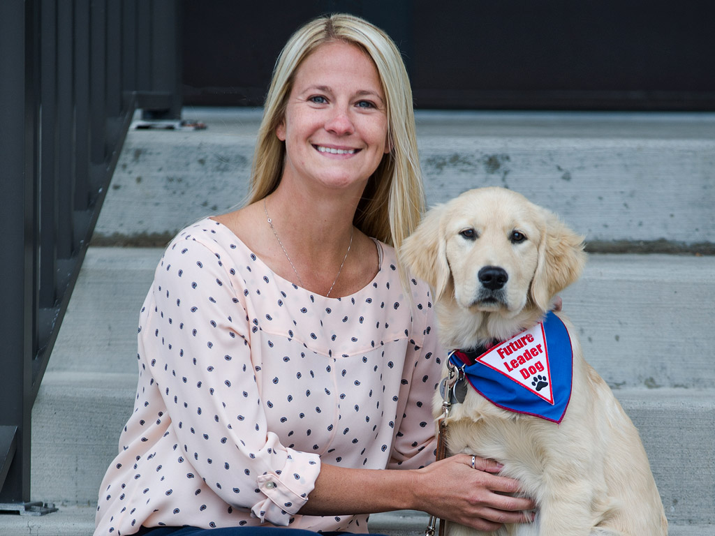 A smiling woman sits on steps beside a golden retriever wearing a blue and red bandana that reads 