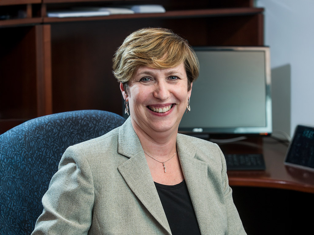 A smiling woman in a light gray blazer sits confidently in an office setting, with a computer visible in the background.