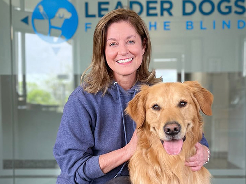 A smiling woman in a hoodie poses with a golden retriever in front of a sign for Leader Dogs for the Blind.