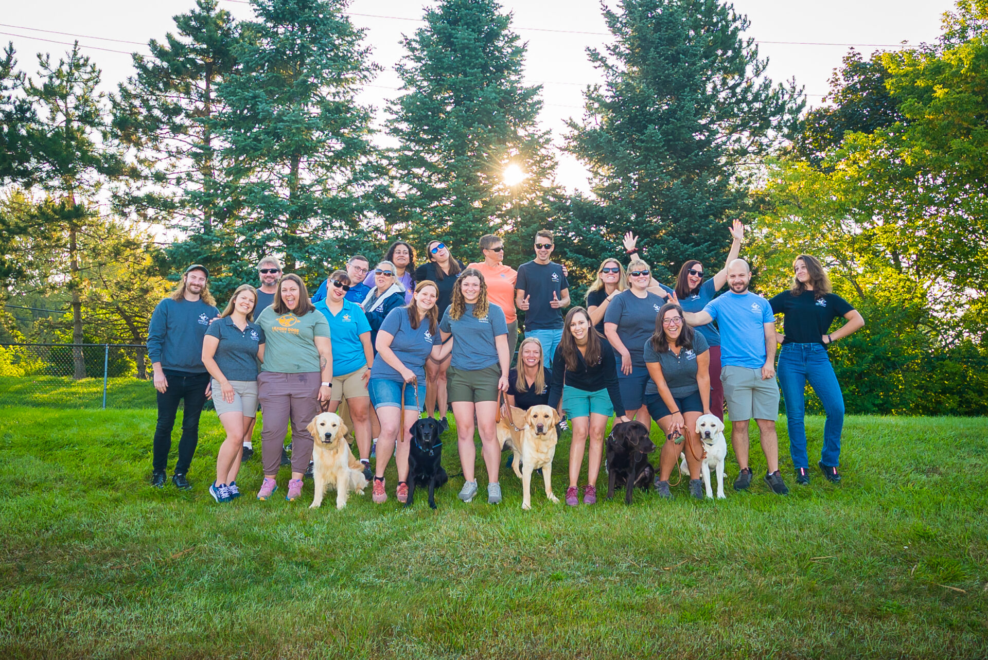 A group of people wearing Leader Dog apparel stand on a grassy hill smiling and showing silly personalities. Four dogs are also seen among the group looking toward the camera.