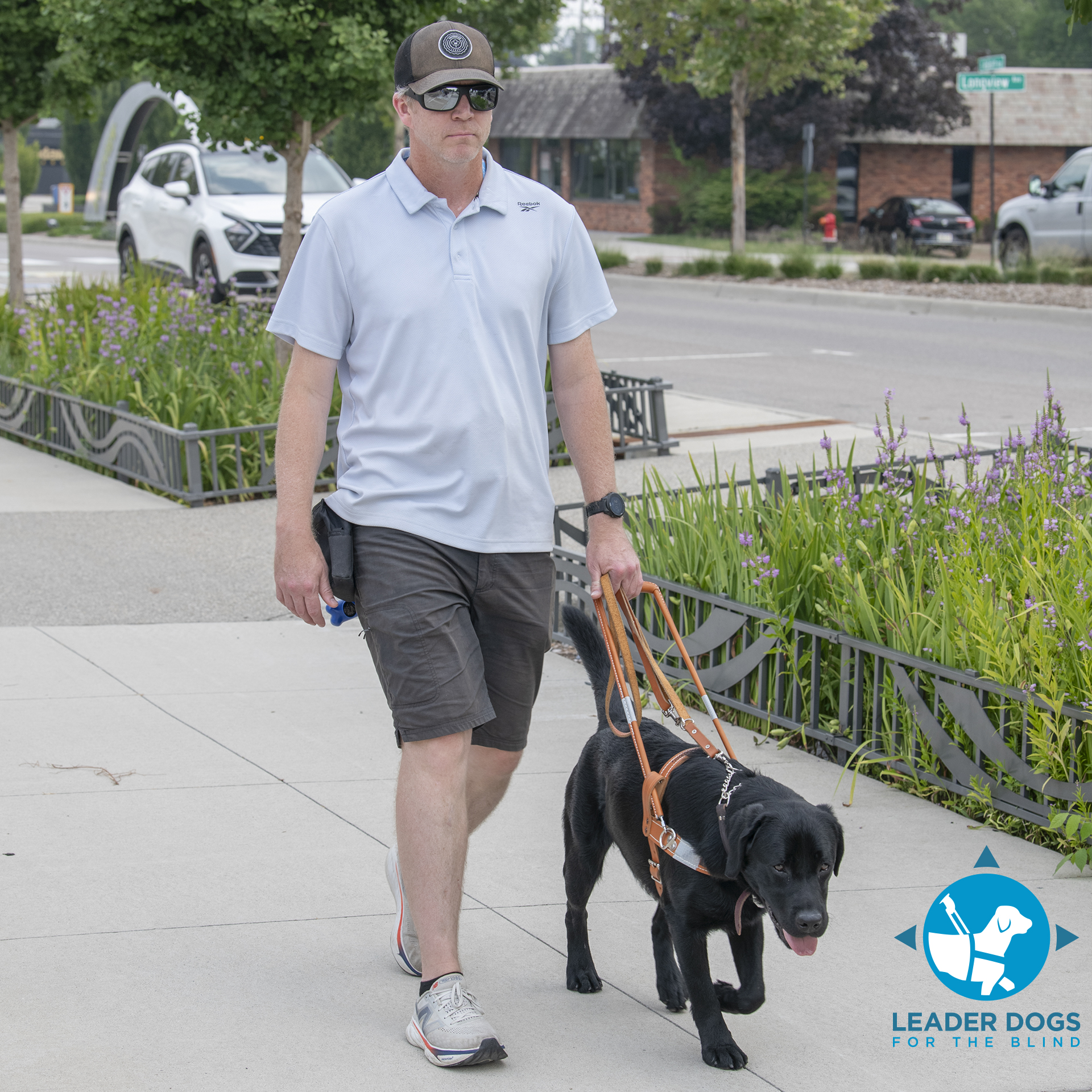 A man walks confidently beside a guide dog, navigating a landscaped urban environment.