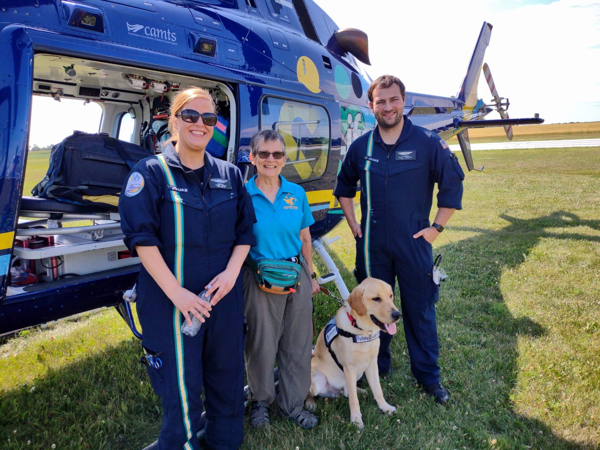 Two medical personnel, a woman and a man, stand alongside a woman and a service dog in front of a colorful helicopter on a grassy field.