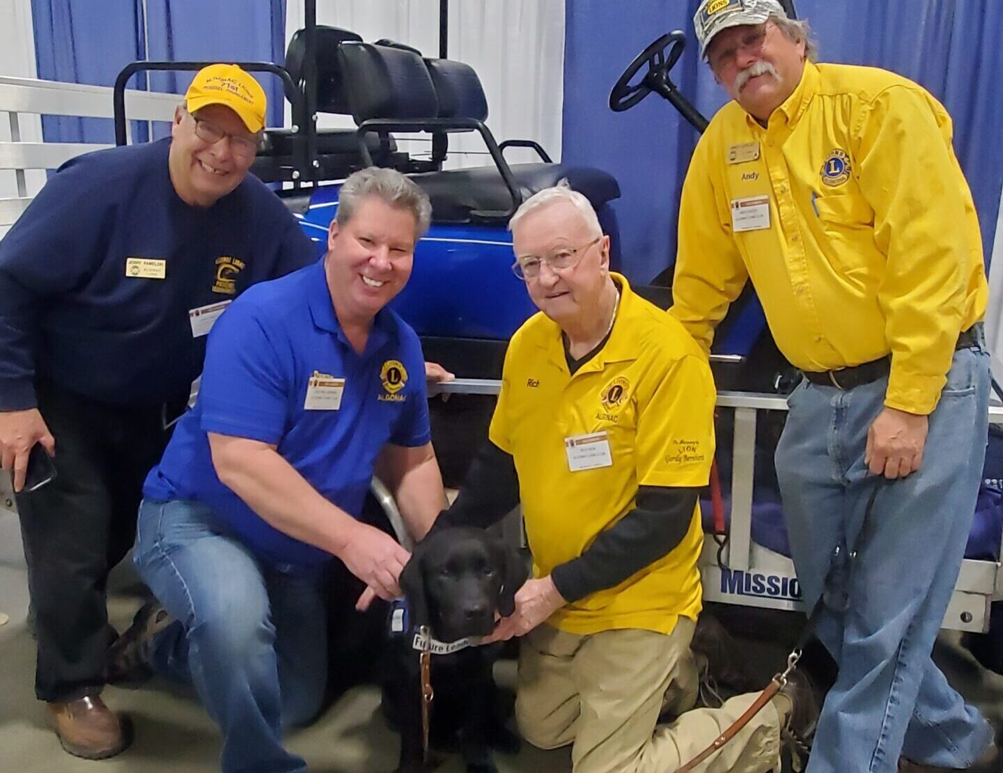 A group of people in matching shirts pose with a service dog beside a golf cart.