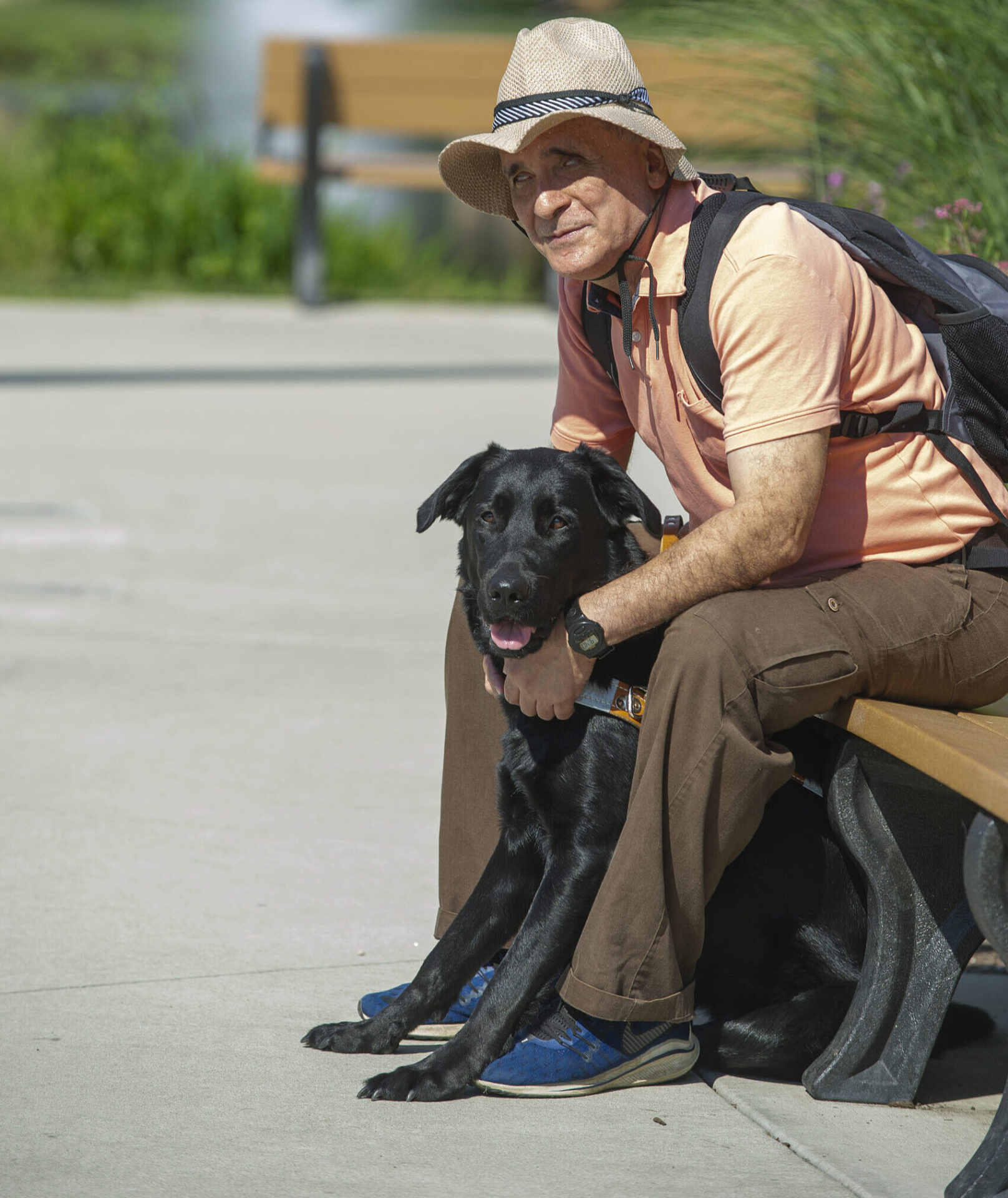 A man wearing a straw hat and light-colored shirt sits on a bench with a black dog beside him, enjoying a sunny day in a park.