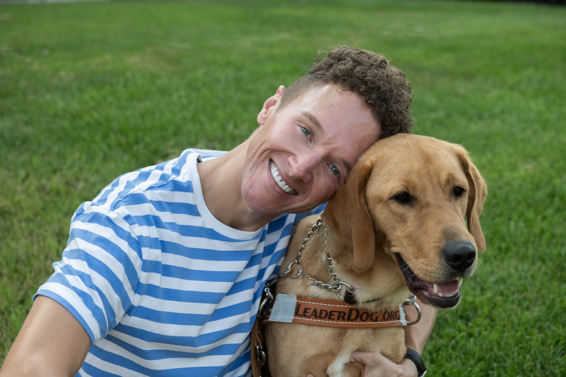 A smiling individual poses affectionately with a Labrador Retriever against a backdrop of green grass.