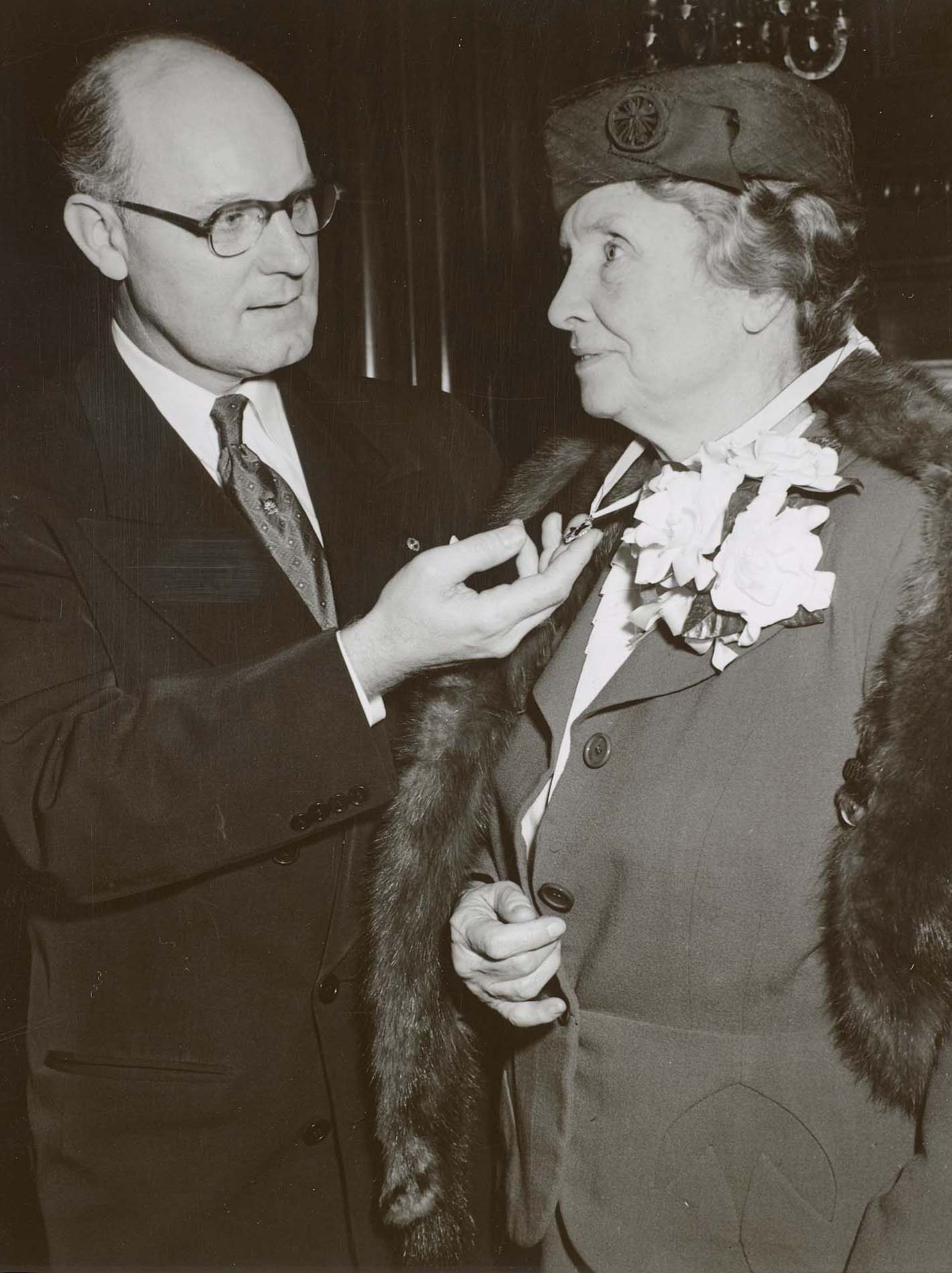 A historic black and white photo of a man and a woman, Helen Keller, as she receives an award pin.