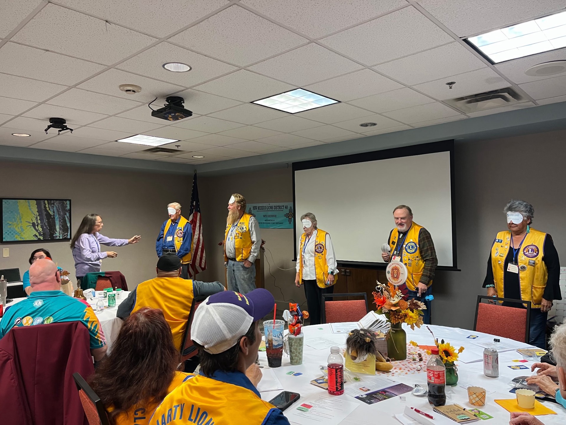 A group of individuals wearing yellow vests stands in a conference room during a meeting, while another person addresses them from the front.