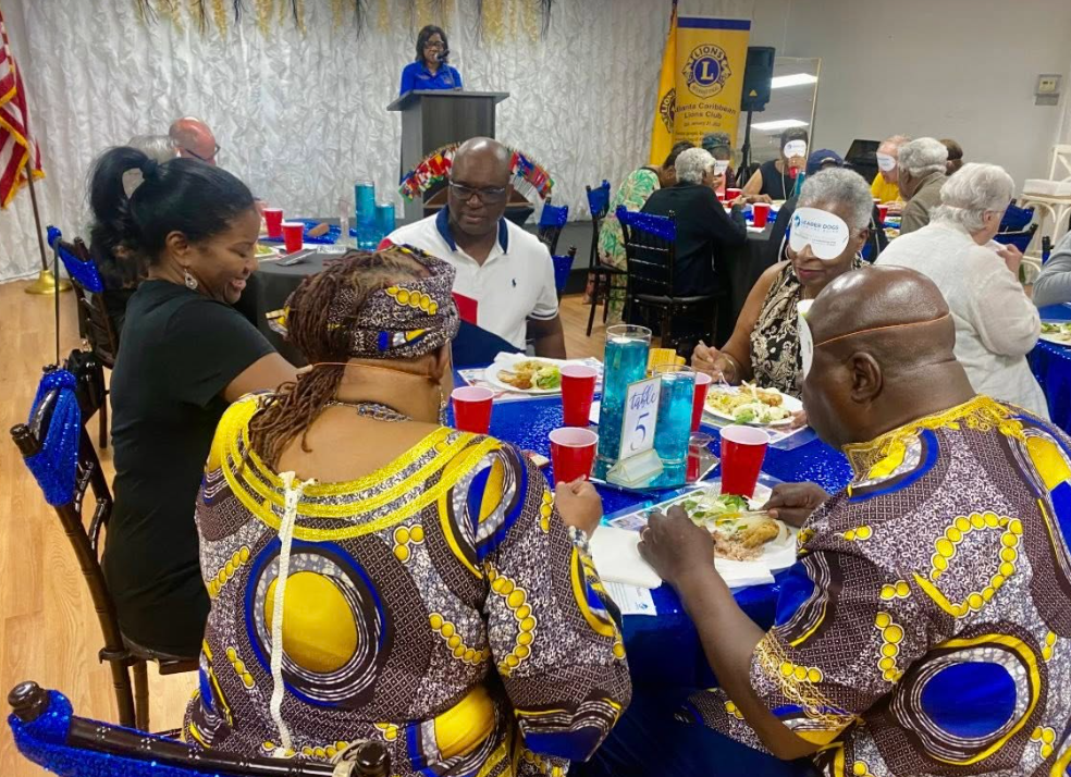 A room full of people wearing blindfolds while eating a Jamaican dinner. A stage can be seen in the back of the room where someone is speaking at a podium.