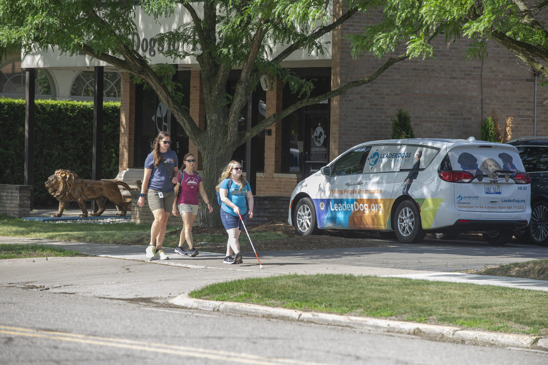 A group walks along a sidewalk near a parked vehicle with a promotional wrap for a guide dog organization, featuring a lion sculpture nearby.