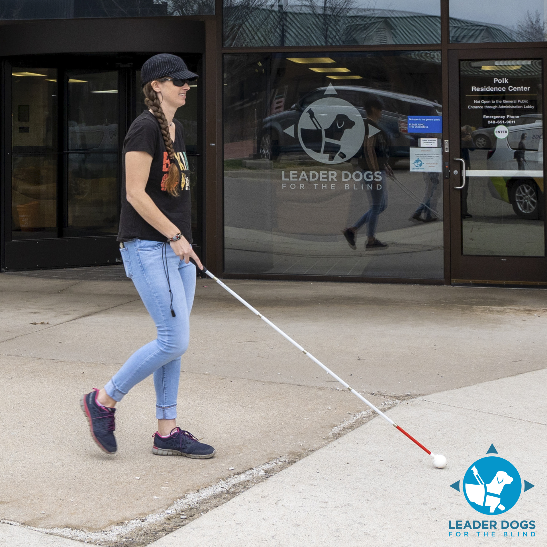 A person with a white cane walks confidently in front of a building with a logo for Leader Dogs for the Blind.