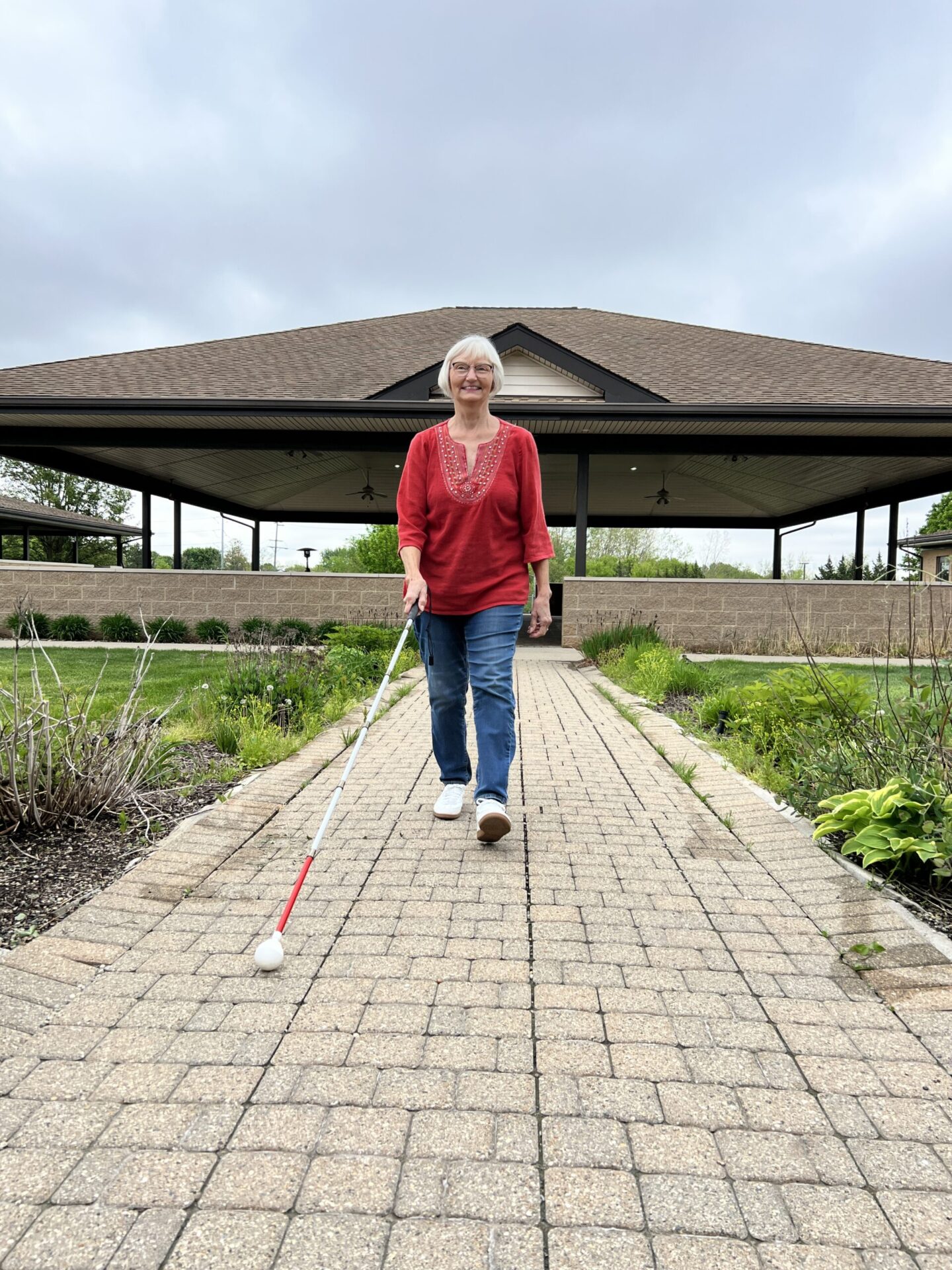 Woman with white cane walking along brick paver pathway away from covered pavilion building.