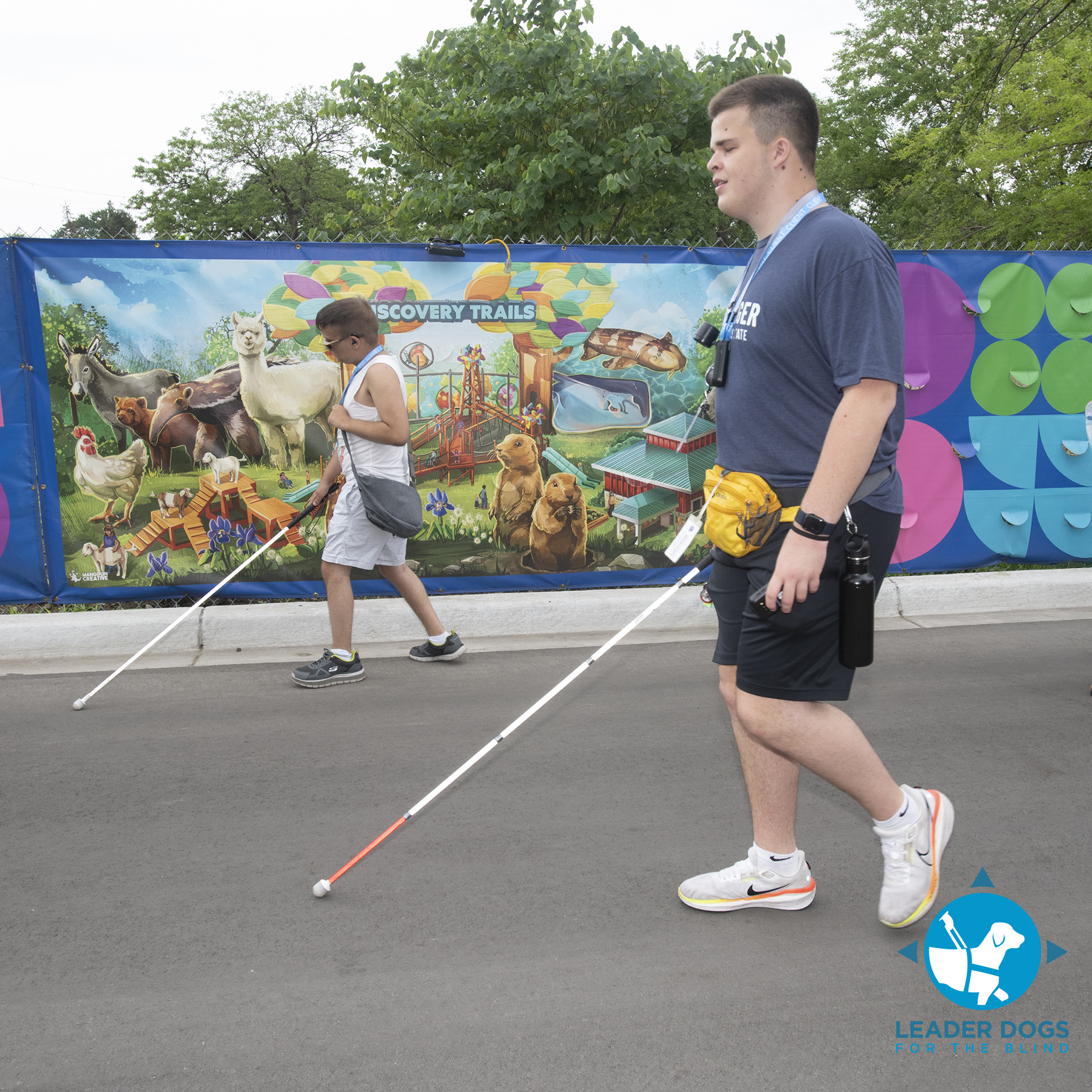 Two individuals are walking along a pathway, using white canes, in front of a colorful mural depicting animals and a vibrant scene.
