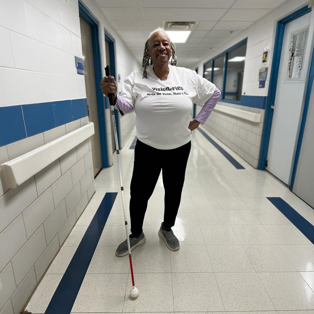 A person stands confidently in a hallway wearing a white t-shirt and holding a white cane.