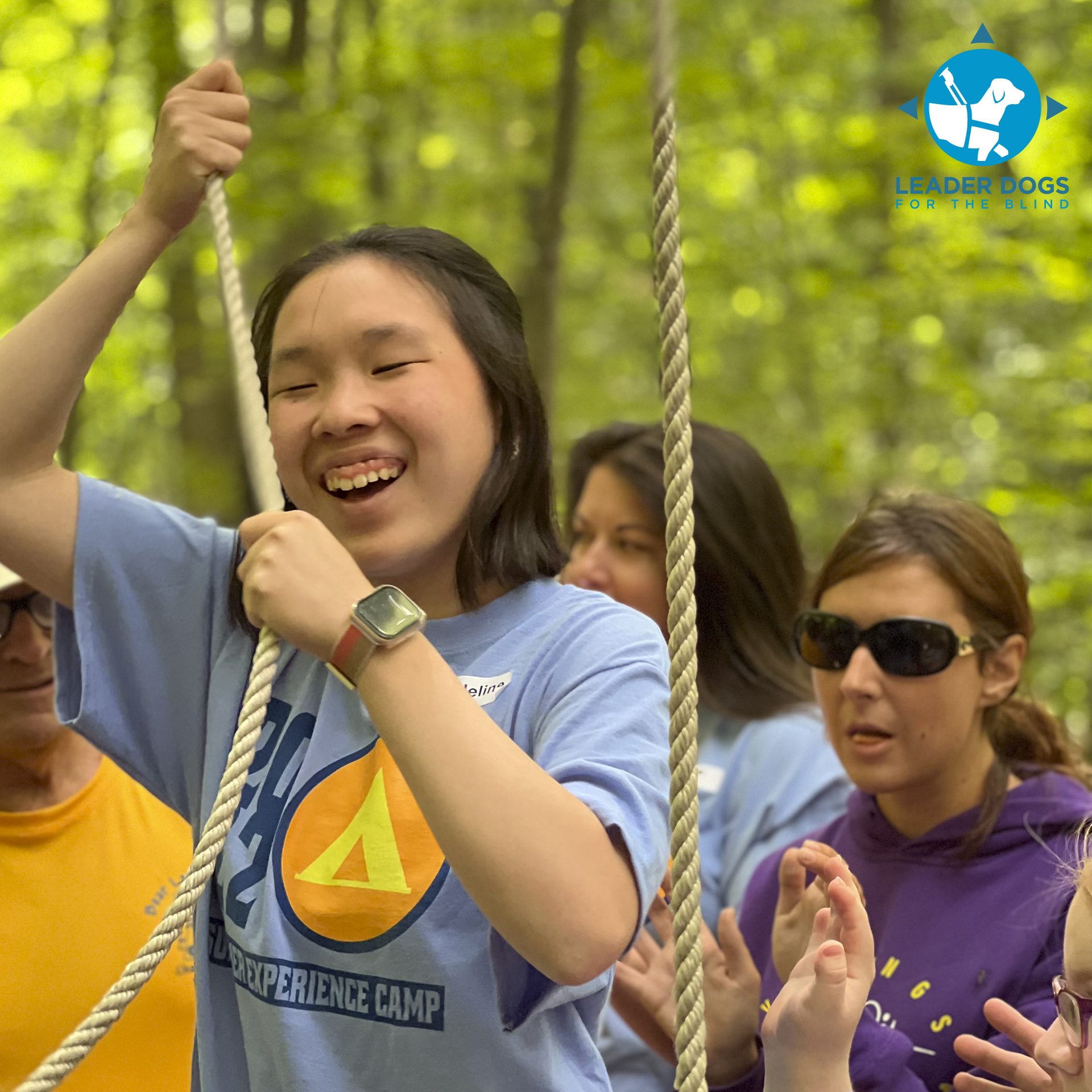 A joyful child pulls on a rope while surrounded by adults, all engaged in a lively outdoor activity amidst lush green trees.