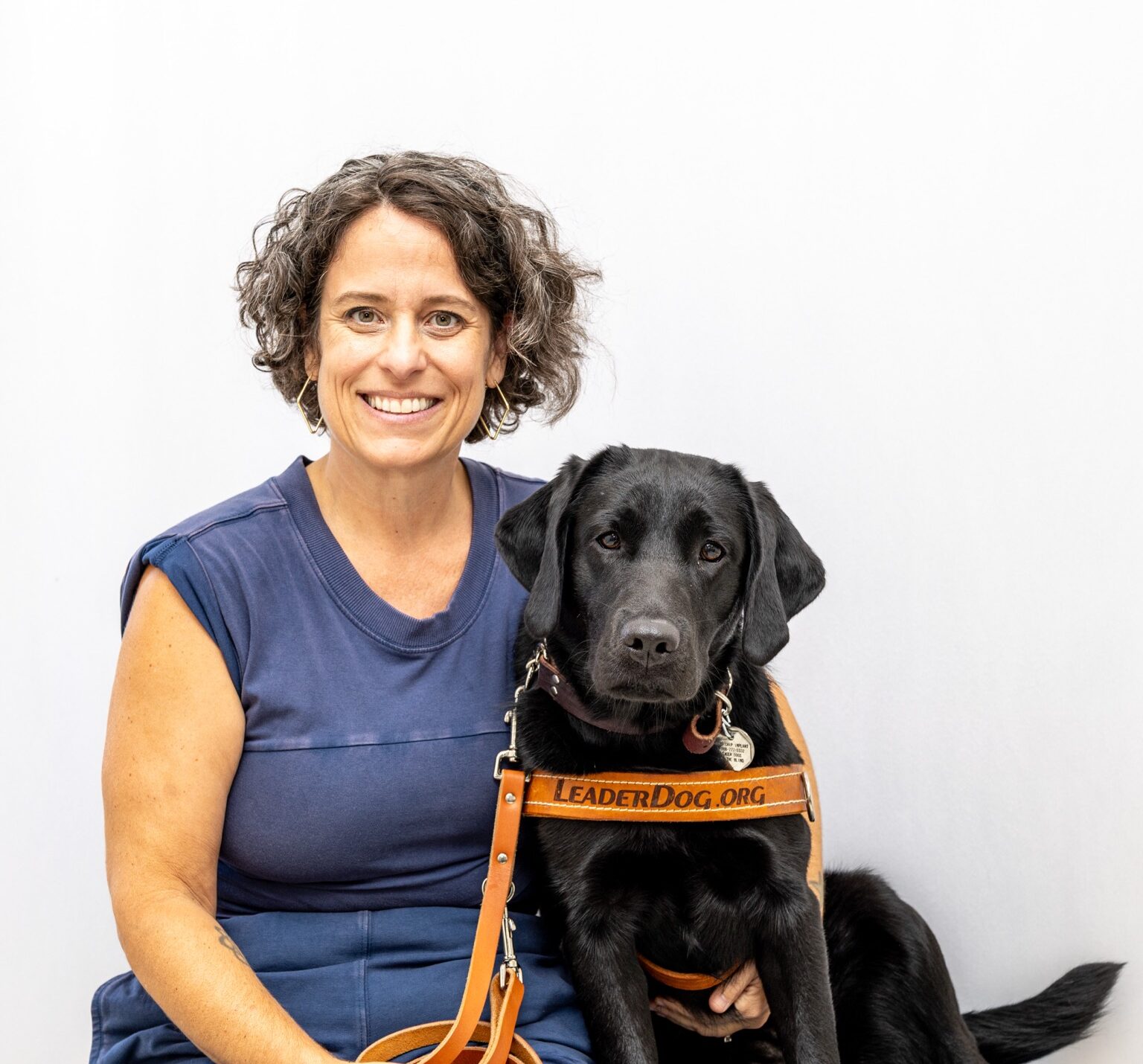 A smiling woman sits next to a black dog wearing a harness that reads 