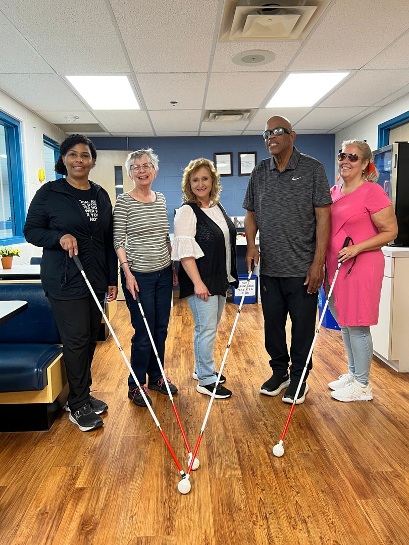 A diverse group of individuals stands together in a bright indoor setting, each holding a white cane and smiling.