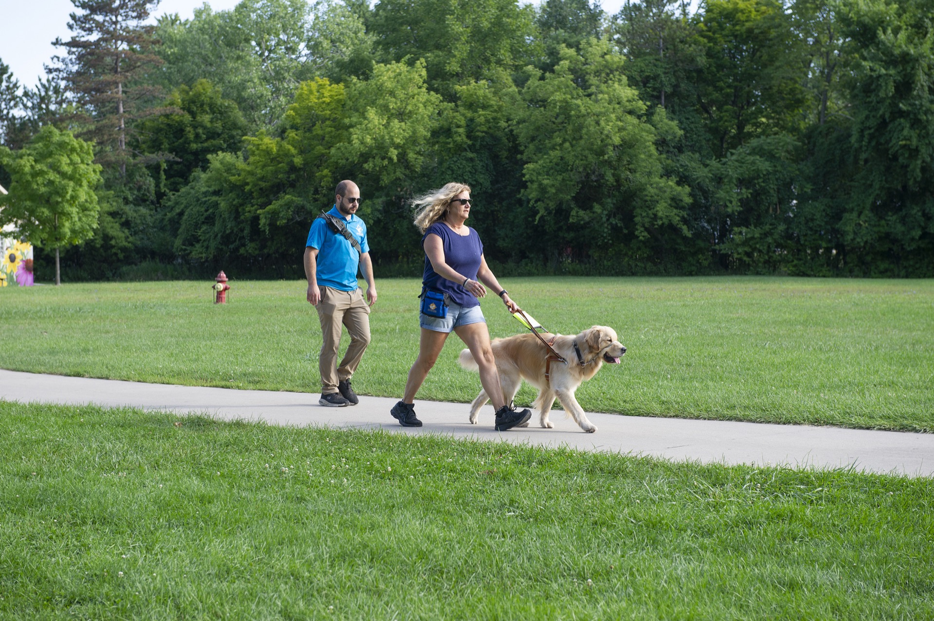 A woman walks a golden retriever along a paved path in a green park while another person follows behind.
