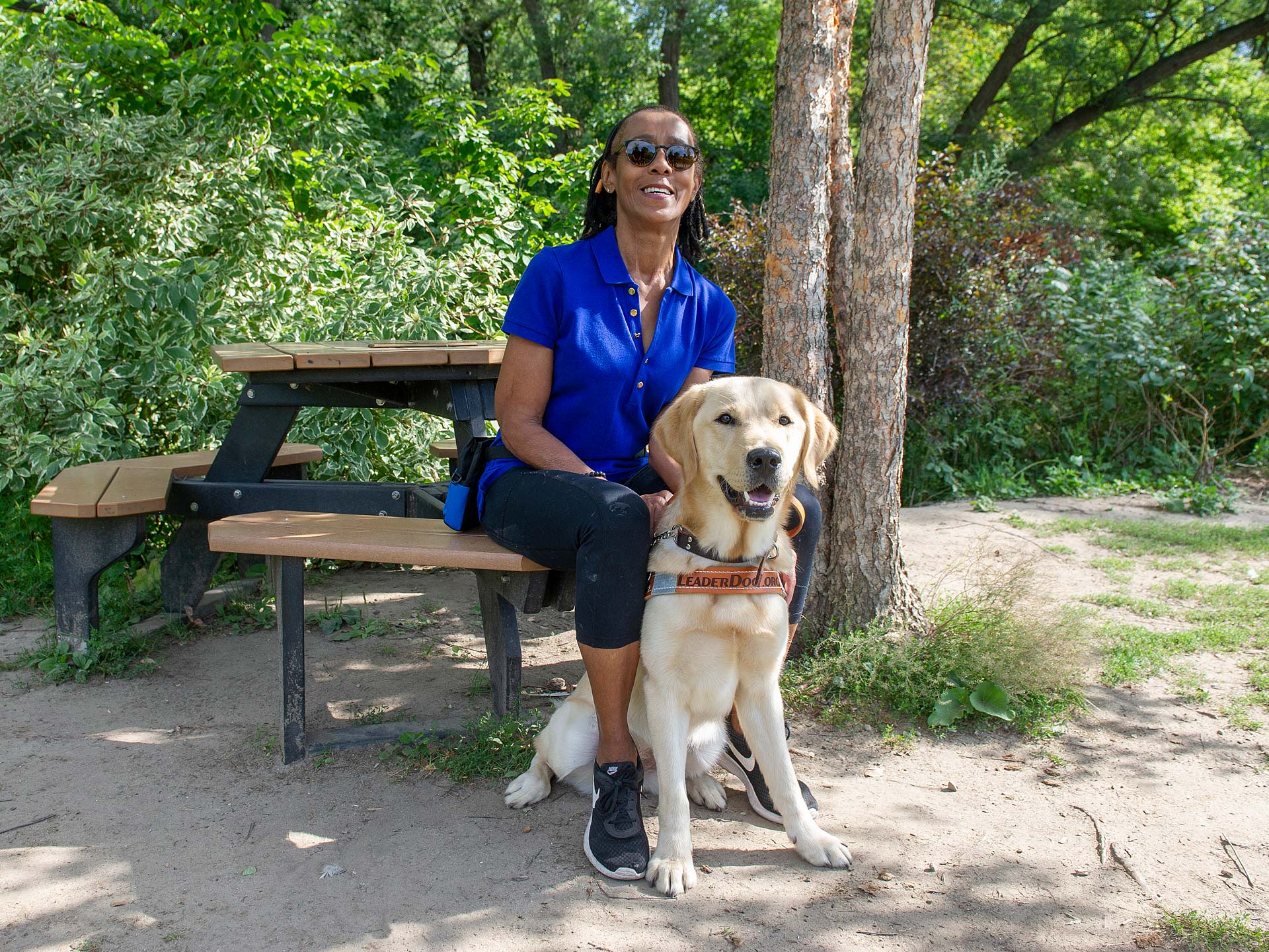 A woman in a blue shirt sits on a bench with a yellow Labrador retriever beside her in a wooded area.