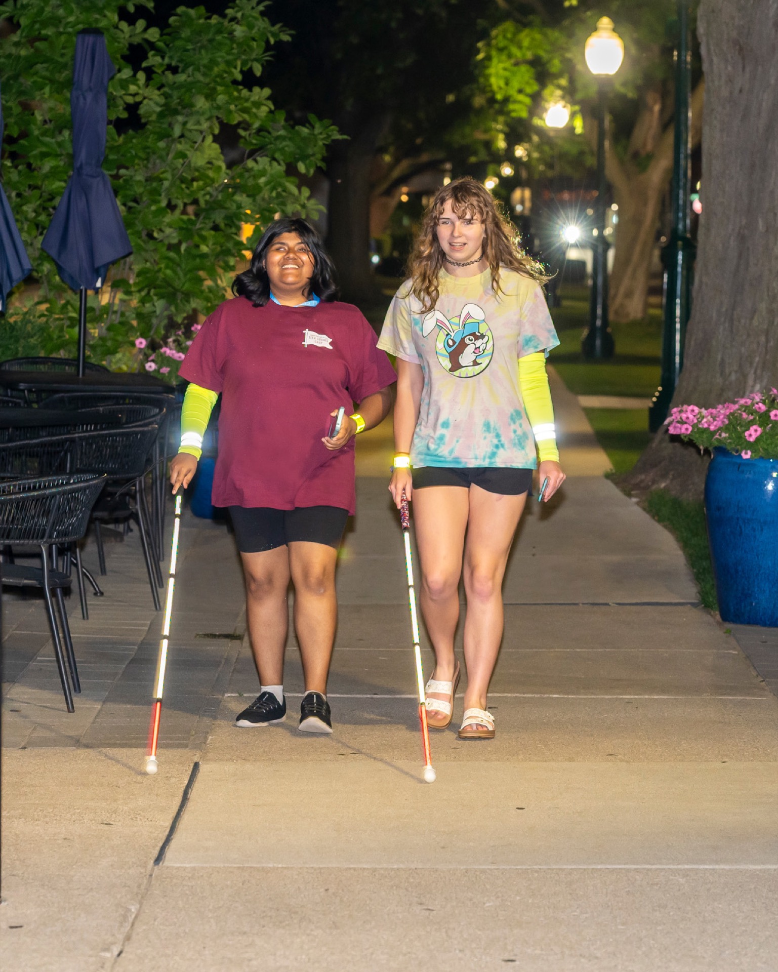 Two individuals are walking together on a well-lit pathway lined with trees and flowers, using canes for navigation.