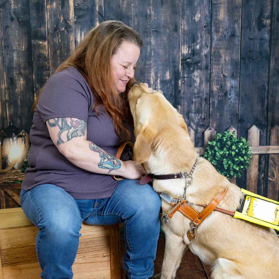 A person smiles as a dog affectionately leans in for a kiss, set against a rustic wooden backdrop.