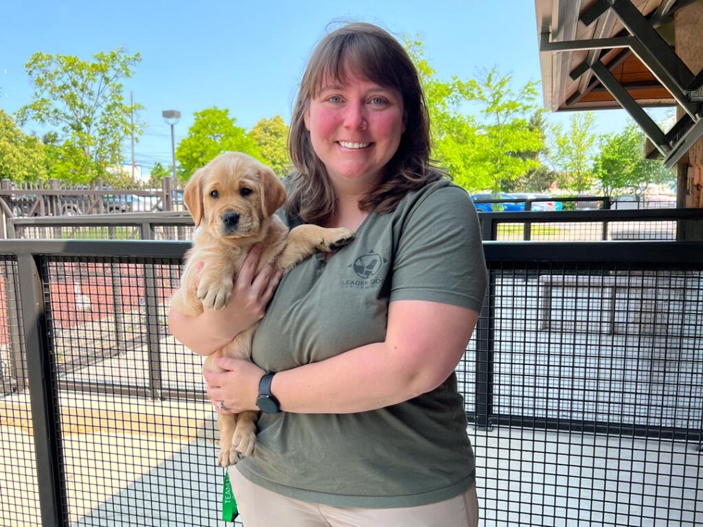 A woman holds a golden retriever puppy while smiling in a sunny outdoor setting.