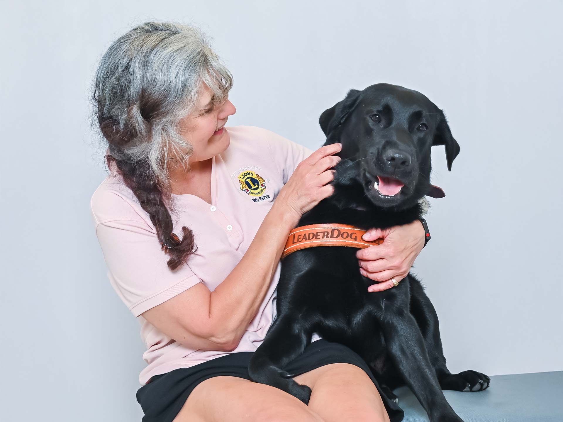 A smiling individual interacts affectionately with a black Labrador wearing a harness labeled 