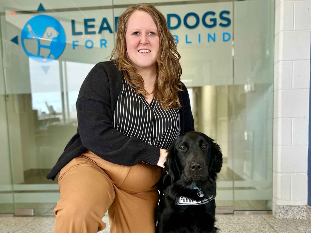A woman in professional attire poses with a black service dog in front of a glass wall displaying the 