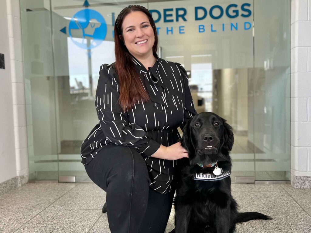 A woman in a black and white patterned blouse kneels beside a black dog wearing a collar that says 