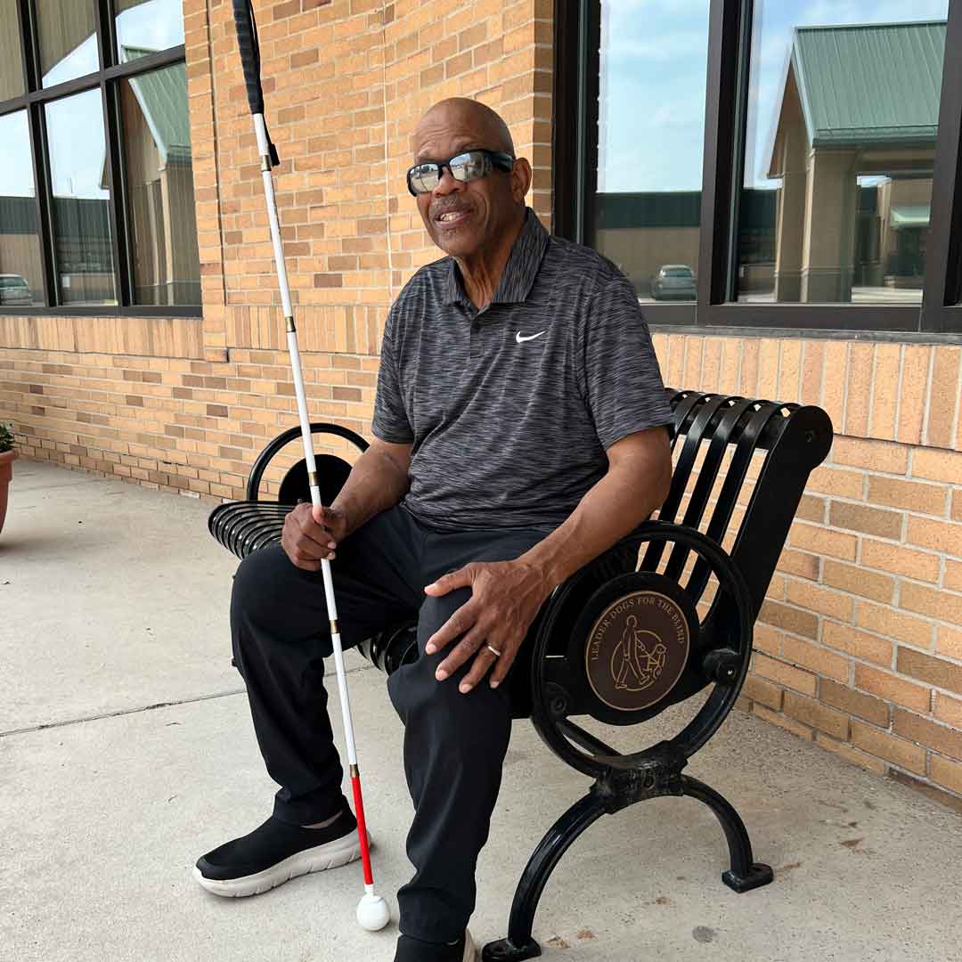 A man wearing glasses sits on a bench outside a building, holding a white cane and smiling.
