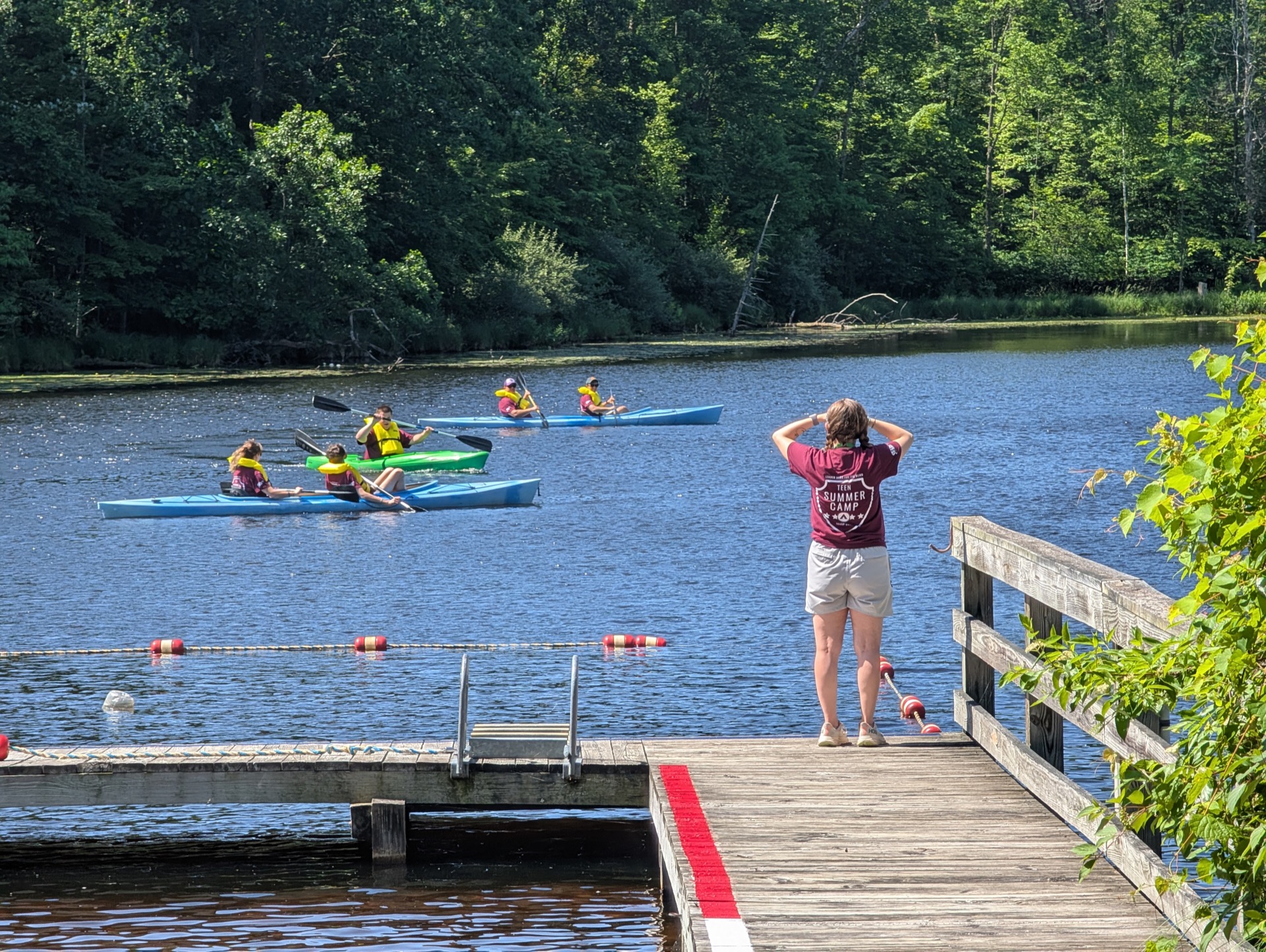 A person stands on a dock, watching kayakers paddling on a calm lake surrounded by lush greenery.