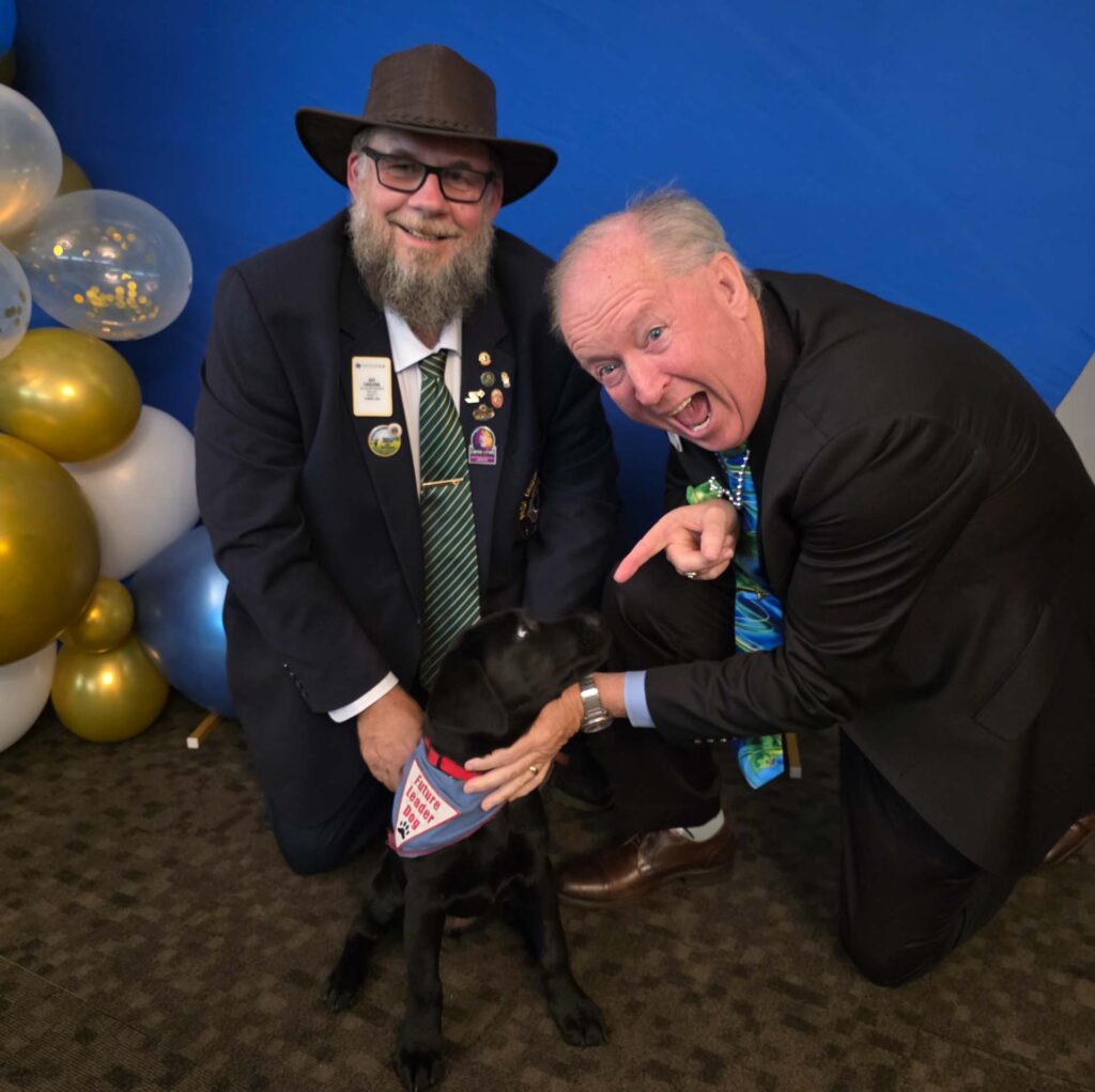 Two men kneels with a black lab puppy in front of a blue backdrop surrounded by balloons. One of the men has a long beard and is wearing a hat, looking toward the camera smiling. The second man is petting the puppy and pointing excitedly at it.
