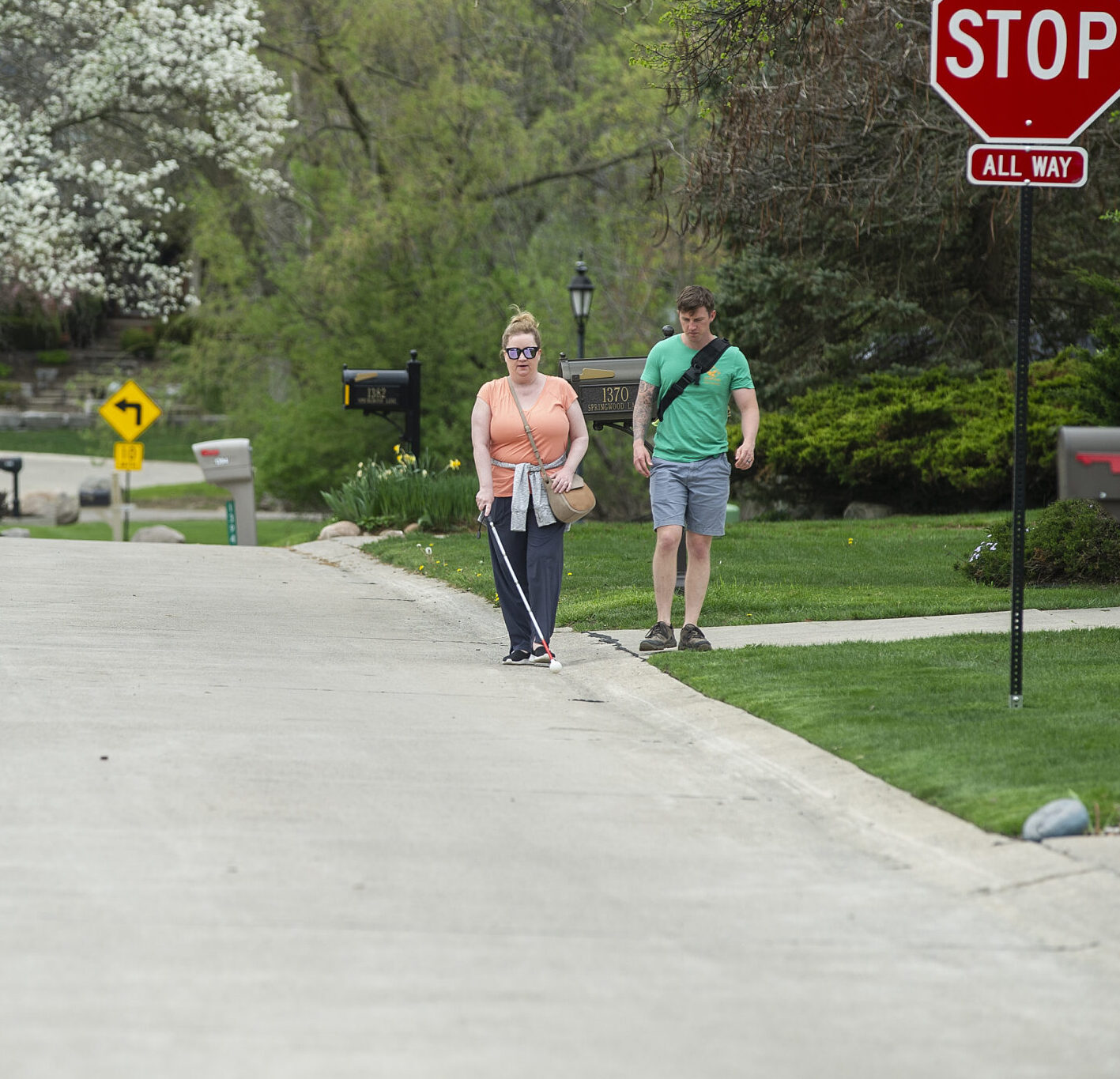 A person with a white cane walks along a pathway guided by another individual in a suburban setting.