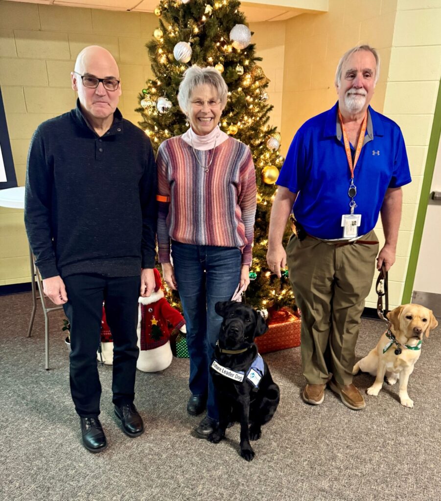 Three adults stand together in front of a Christmas tree, two men and one woman. The woman stands with a black lab wearing a Future Leader Dog vest, to her rights is a man holding the leash of a yellow lab wearing a Leader Dog canine ambassador vest. All three adults smile toward the camera.