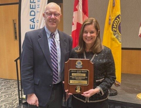 A man and woman smile toward the camera while standing in front of a stage. The woman is holding a wooden plaque.