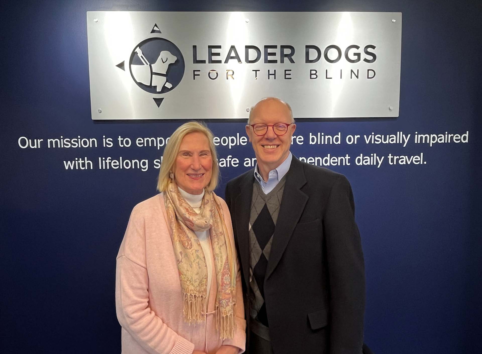 A woman and a man smile toward the camera standing in front of a Leader Dogs for the Blind sign.