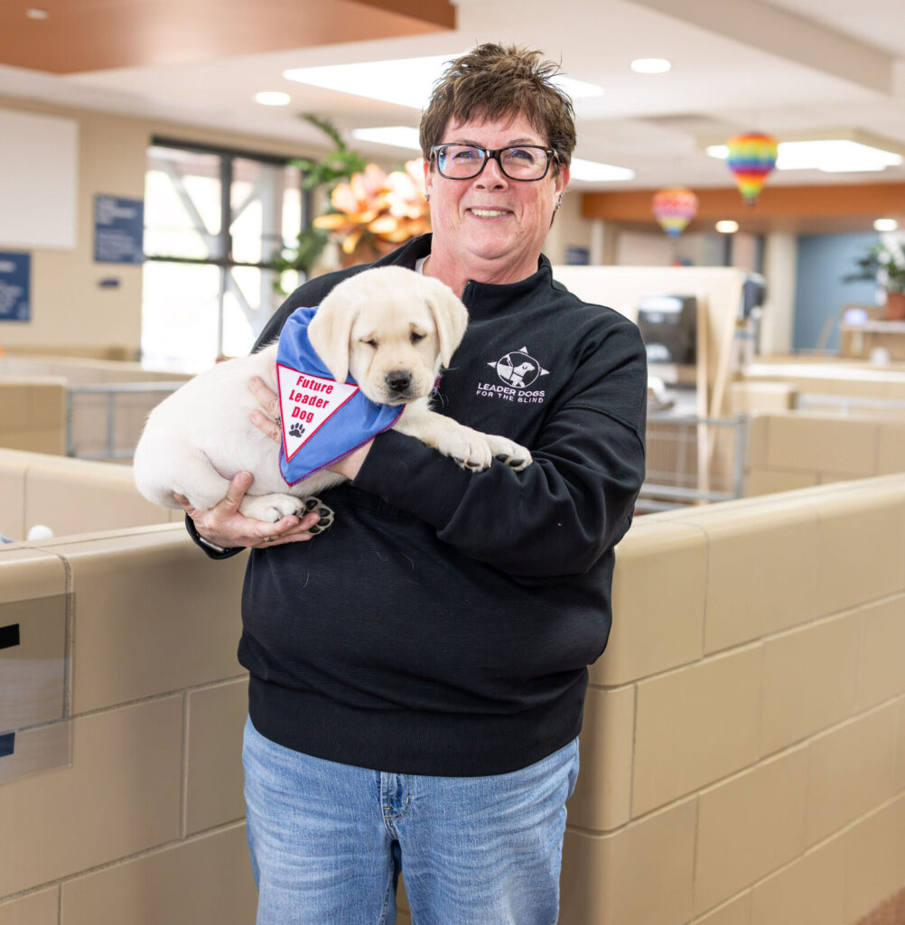 A woman with short hair and glasses stands smiling, holding a yellow lab puppy that is wearing a 