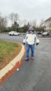 A person walks on the side of the road along the curb using a long white cane. The person is dressed in a large winter coat.