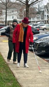 Two people walk along a sidewalk on a cold day. One person walks ahead using a long white cane, while another person walks behind smiling toward the camera.