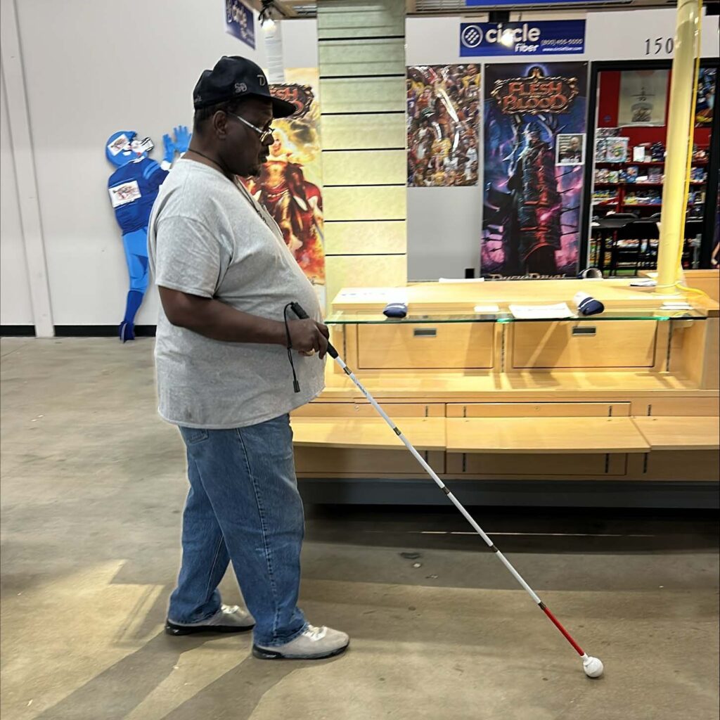 A man uses his white cane as he walks past a kiosk in the antique mall in which he runs a store. He is wearing a dark baseball cap, a gray t-shirt, and jeans.