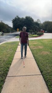 A man uses his white cane as he travels on the sidewalk. He is wearing a plaid shirt and blue jeans. In the background are crepe myrtles.