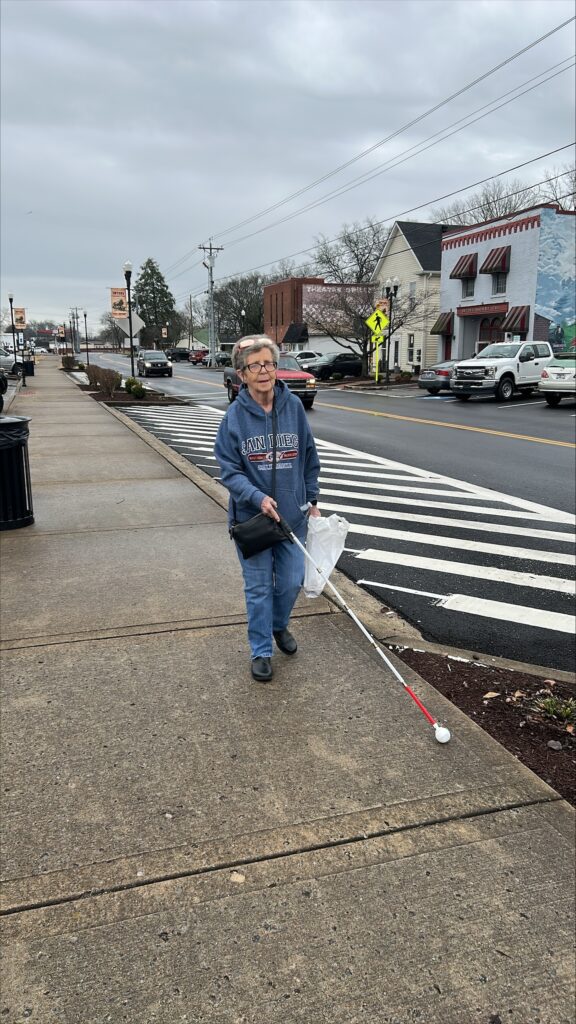 A woman walks with a long white cane along a wide sidewalk in a small downtown area. She is holding a white plastic bag.