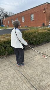 A woman walks along a sidewalk near a brick building using a long white cane.