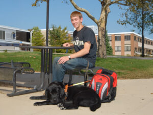 A young man sits at a table outdoors with a black dog resting at his feet beside a red backpack.