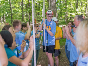 A group of people in matching blue shirts enthusiastically supports someone navigating a rope swing in a green forest setting.