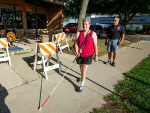 A person is walking along a sidewalk using a white cane, while another individual stands nearby, observing the situation.
