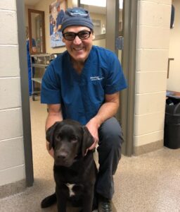 A man wearing surgical scrubs and cap kneels behind a black lab.