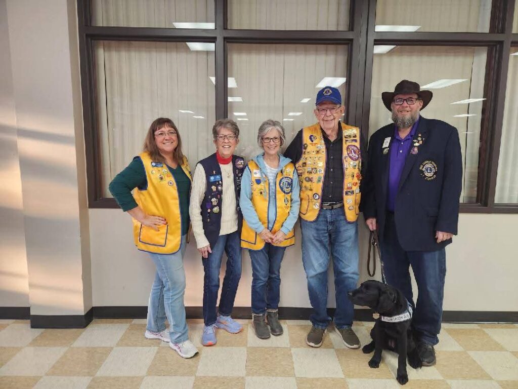 Four adults stand together smiling toward the camera. They all wear Lions Club International vests or jacket covered in pins. The black lab wears a Future Leader Dog vest.
